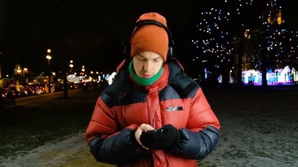 Молодой кавказский мужчина слушает музыку в наушниках на вашем телефоне, танцует и поет от радости, глаза закрыты, наслаждаясь музыкой в городе сегодня вечером гуляя зимой . — стоковое видео