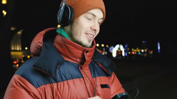 Joven hombre caucásico escuchando música en auriculares en su teléfono, bailando y cantando de alegría, los ojos cerrados, disfrutando de la música en la ciudad esta noche caminando en el invierno . — Vídeo de stock