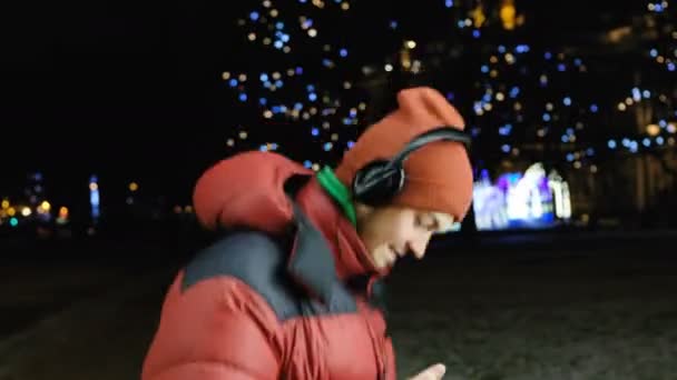 Kaukasische Young man, luisteren naar muziek op de hoofdtelefoon op uw telefoon, dansen en zingen van vreugde, ogen dicht, genieten van de muziek in de stad vanavond wandelen in de winter. — Stockvideo