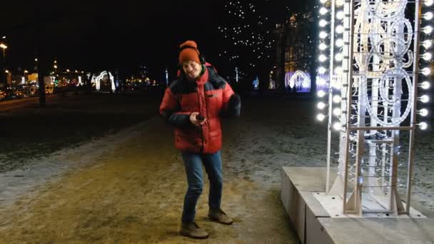 Joven hombre caucásico escuchando música en auriculares en su teléfono, bailando y cantando de alegría, los ojos cerrados, disfrutando de la música en la ciudad esta noche caminando en el invierno . — Vídeo de stock