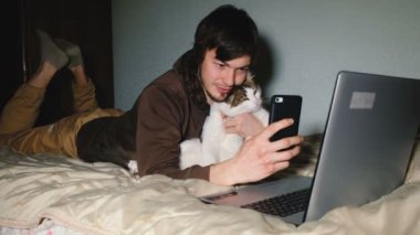Beyaz delikanlı smartphone yatak laptop önünde yatan bir selfie kedi ile taking.