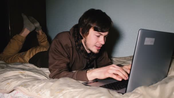 ベッドと彼女の友人のノート パソコンとテキスト メッセージで横になっている若い男. — ストック動画