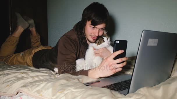 Jovem caucasiano tomando uma selfie com o gato no smartphone deitado na cama na frente do laptop . — Vídeo de Stock