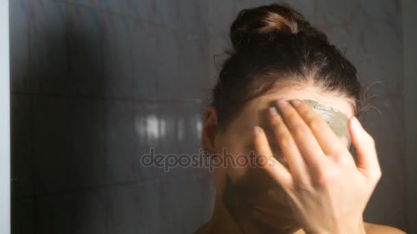 Menina sorrindo jovem aplicando uma máscara cosmética de barro azul no banheiro, 4K . — Vídeo de Stock