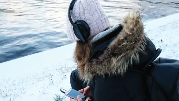 Дівчина слухає музику на навушниках на березі річки взимку, 4k . — стокове відео