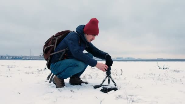 El hombre pone un trípode con un teléfono inteligente en el suelo de nieve, un trípode para el teléfono, 4K . — Vídeo de stock