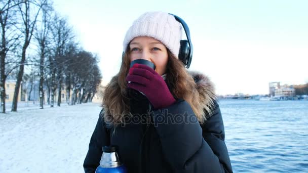 Menina bonita ouve música e bebe chá de uma garrafa térmica ao ar livre no inverno, aquece no parque, 4k . — Vídeo de Stock