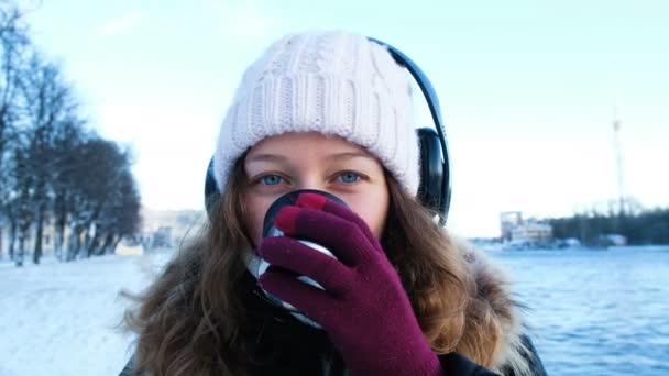 Vacker ung flicka tittar på kameran, lyssnar på musik och dricka te från en termos utomhus på vintern, värmde i parken, porträtt av en flicka närbild, 4k. — Stockvideo