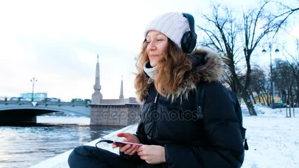 Jong meisje luistert naar muziek buiten in de winter, zingt het liedje en dansen zittend op de sneeuw, 4k. — Stockvideo