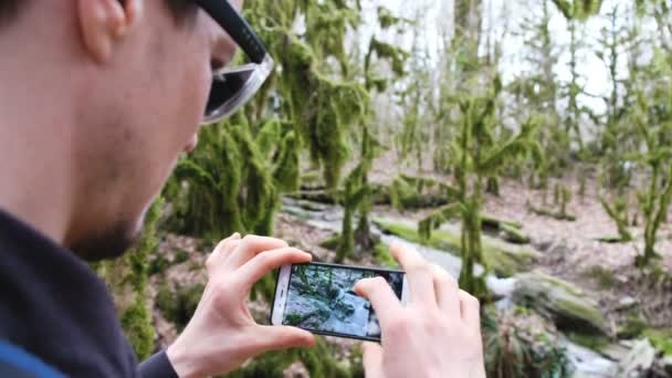 A man in glasses photographs a stream in the forest on a smartphone, 4k. — Stock Video