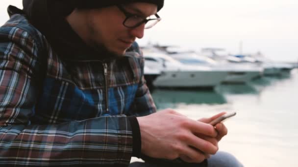 Ένας άνθρωπος κάθεται στο εδώλιο του κατηγορουμένου στη θάλασσα και χρησιμοποιεί ένα smartphone, ένας άνθρωπος που κάθεται στο διαδίκτυο από το dock, 4k. — Αρχείο Βίντεο