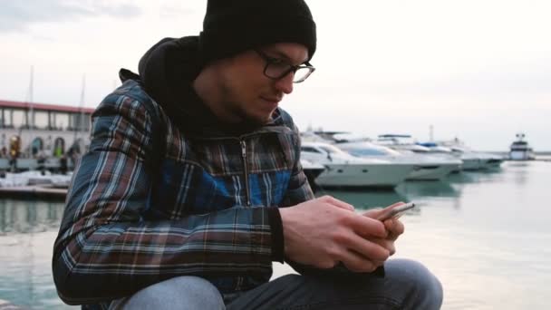 El hombre se sienta en el muelle del mar y utiliza un teléfono inteligente, 4k . — Vídeos de Stock