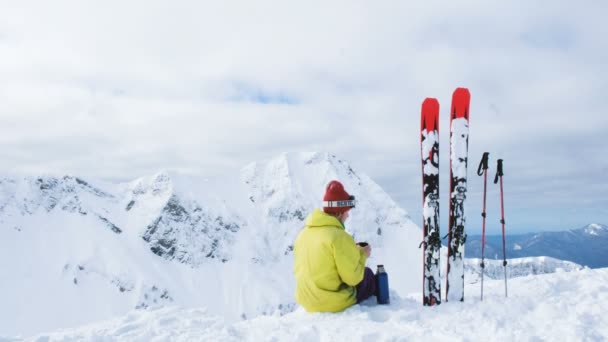 Skier in a bright suit sitting on top of a mountain and drinking tea, there are a number of skis, 4 K. — Stock Video