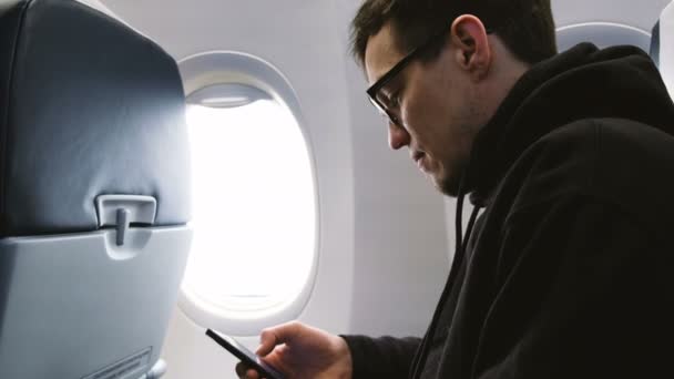 Ο άνθρωπος με τα γυαλιά χρησιμοποιεί ένα smartphone σε ένα αεροπλάνο, κάθεται στο παράθυρο, 4k. — Αρχείο Βίντεο