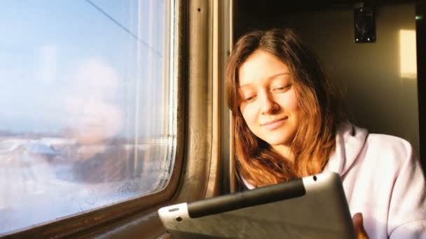Menina caucasiana bonita goza de um tablet no trem, sorrindo e olha pela janela ao pôr do sol, 4k . — Vídeo de Stock