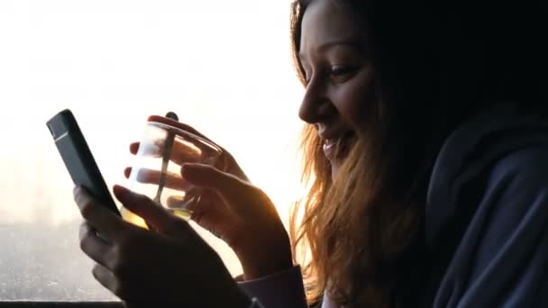 Όμορφο κορίτσι Καυκάσιος είναι πόσιμο τσάι και χρησιμοποιώντας ένα smartphone σε φόντο ηλιοβασίλεμα σε ένα τρένο, 4k. — Αρχείο Βίντεο