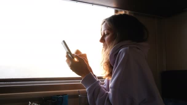 Schöne kaukasische Mädchen verwendet ein Smartphone auf einem Hintergrund von Sonnenuntergang Strahlen in einem Zug, 4k. — Stockvideo