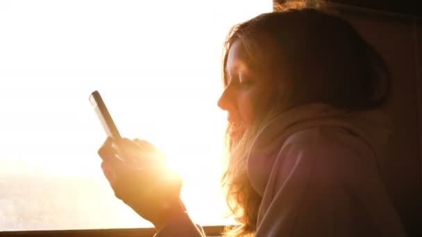 Hermosa chica caucásica imprime un mensaje en un teléfono inteligente sobre un fondo de puesta de sol en un tren, 4k . — Vídeo de stock