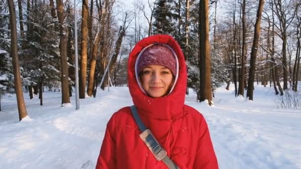 En flicka i en röd dunjacka promenader och ser rakt in i kameran i skogen på vintern vid solnedgången, Slowmotion — Stockvideo