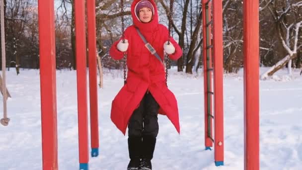 Una ragazza adulta con un piumino rosso che oscilla su un'altalena rossa nel parco giochi al tramonto, al rallentatore — Video Stock