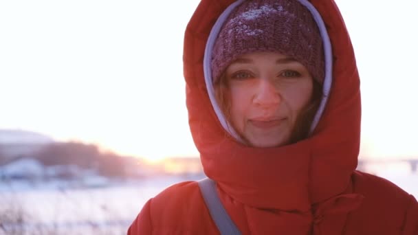 Una ragazza con un piumino rosso guarda direttamente nella fotocamera in inverno sullo sfondo di un declino, rallentatore — Video Stock