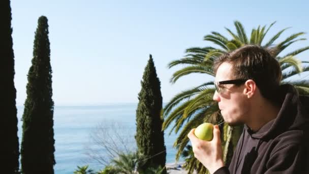 El hombre está comiendo una manzana sobre un fondo de palmas y el mar — Vídeos de Stock