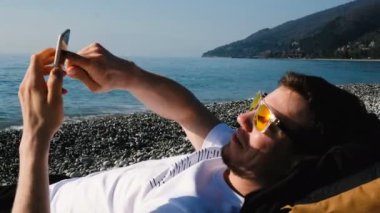 Adam sahilde deniz güneş gözlüğü takıyor ve bir telefon kullanarak dinleniyor