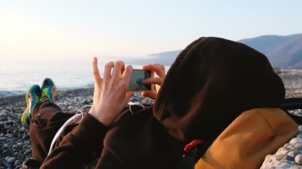 Man is het fotograferen van zijn voeten tegen de achtergrond van de zee bij zonsondergang — Stockvideo