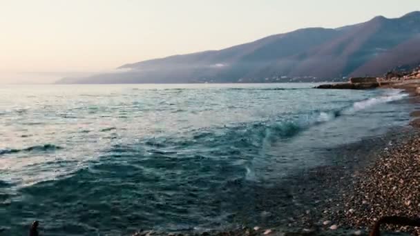 Vlny beat na oblázky, azure moře při západu slunce na pozadí kopců — Stock video