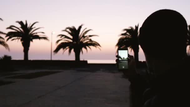 Un hombre está tomando fotos de un panorama de palmeras en el mar al atardecer — Vídeos de Stock