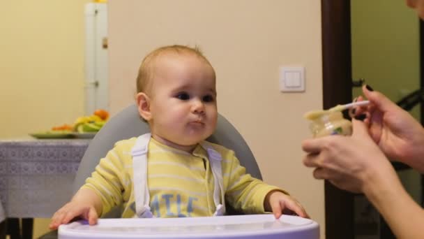 Μωρό κορίτσι τρώει πουρέ από ένα βάζο που κάθεται σε μια καρέκλα παιδική, μαμά δίνει άγευστη μωρό τροφίμων στο σπίτι — Αρχείο Βίντεο
