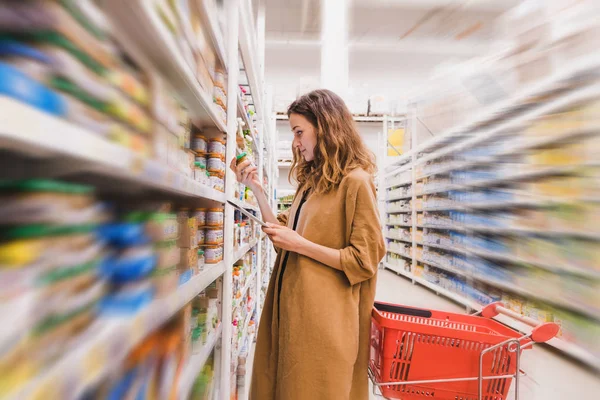 Jovem mulher bonita com um tablet escolhe comida de bebê em um supermercado, fundo borrado — Fotografia de Stock