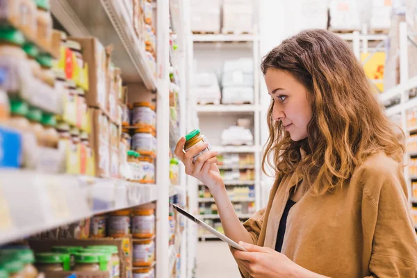 Jovem mulher bonita com um tablet pega comida de bebê em um supermercado, a menina está estudando a composição do produto close-up — Fotografia de Stock