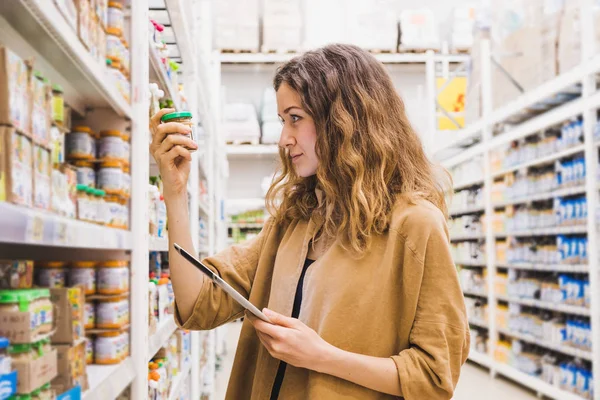 Jovem mulher bonita com um tablet seleciona comida de bebê em um supermercado, a menina lê cuidadosamente a composição do produto. — Fotografia de Stock