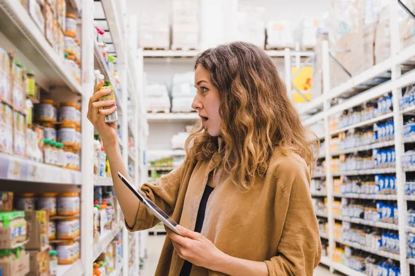 Junge Frau mit Tablette schockiert von der Zusammensetzung der Babynahrung in einem Supermarkt, liest das Mädchen emotional die Beschreibung der Waren — Stockfoto