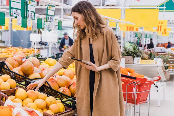 Ung kvinna med en tablett plockar frukt i en stormarknad — Stockfoto