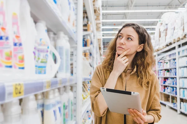 Business nainen ajattelee ostoksia tabletilla valitsee kotitalouksien kemikaaleja supermarketissa — kuvapankkivalokuva