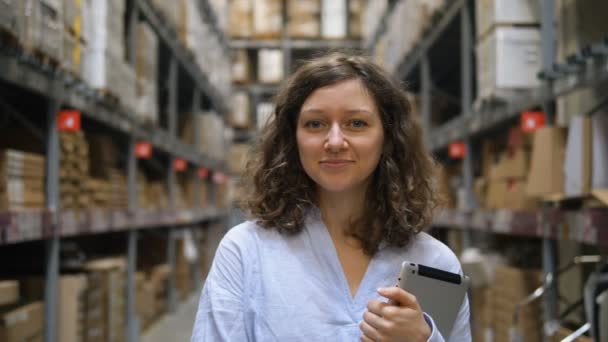 Привлекательная девушка стоит с планшетом на складе между полками с товаром, улыбается и смотрит в камеру — стоковое видео