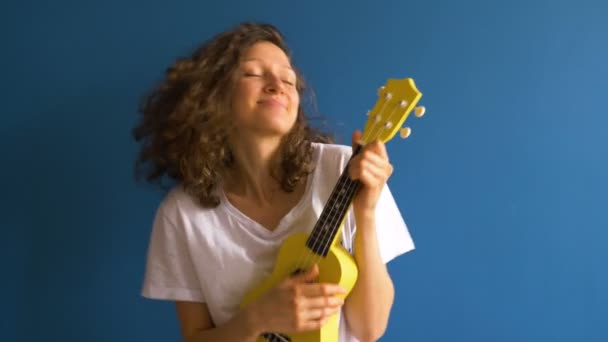 Счастливая молодая женщина играет на жёлтой укулеле на фоне синей стены — стоковое видео