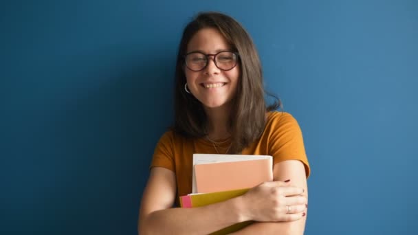 A beautiful woman designer in glasses smiles and looks at the camera holding a bundle of projects in her hands on a blue background — Stock Video