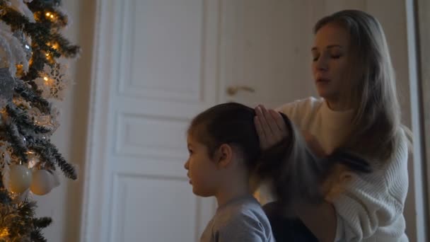 Hübsche Blondine kämmt junge Tochter Haare in der Nähe des Weihnachtsbaums — Stockvideo