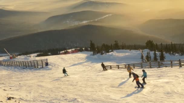 Μια ομάδα σκιέρ και snowboarders κατεβαίνουν την πλαγιά του σκι με φόντο ένα εκπληκτικό ηλιοβασίλεμα και λόφους του βουνού — Αρχείο Βίντεο