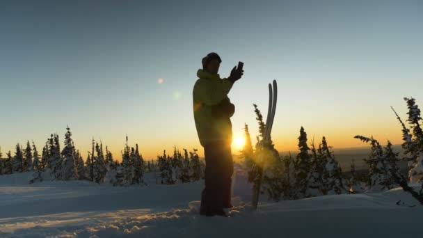 Yetenekli kayakçı akıllı telefonu elinde tutuyor ve gün batımını fotoğraflıyor. — Stok video