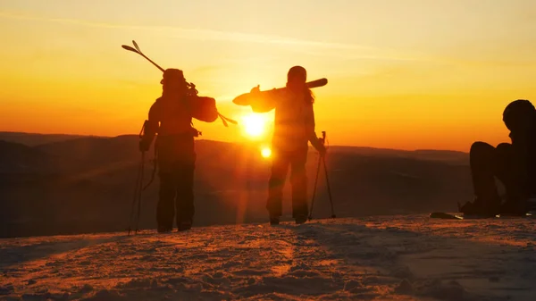 Pessoas com equipamento de esqui caminham ao longo da encosta nevada ao pôr do sol — Fotografia de Stock