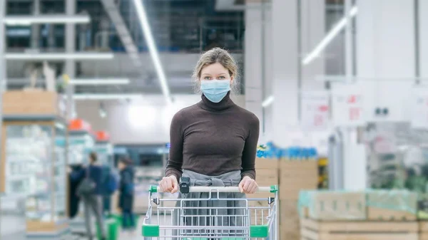Een vrouw met een medisch masker staat in een supermarkt met een tijdloze boodschappenkar. Bescherming tegen het coronavirus, kopen van voedsel in een crisis. — Stockfoto