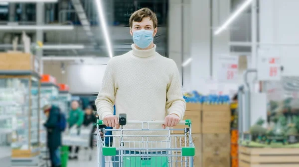 Un uomo con una maschera medica si trova in un supermercato con un timelaps carrello del cibo — Foto Stock