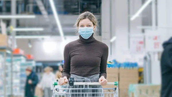 Een vrouw met een medisch masker staat in een supermarkt met een boodschappenkar. Bescherming tegen het coronavirus, kopen van voedsel in een crisis. — Stockfoto