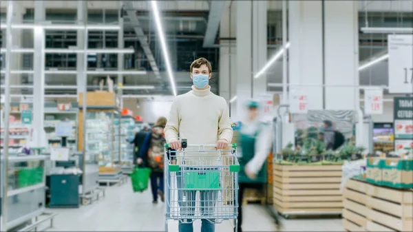 Un uomo con una maschera medica si trova in un supermercato con un carrello del cibo — Foto Stock