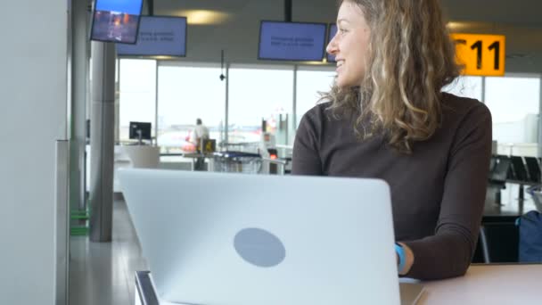 Chica sonriente trabaja en el portátil sentado en el aeropuerto salón cafetería — Vídeos de Stock