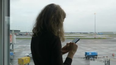kız havaalanının penceresinin yanında cep telefonuyla internette sörf yapıyor